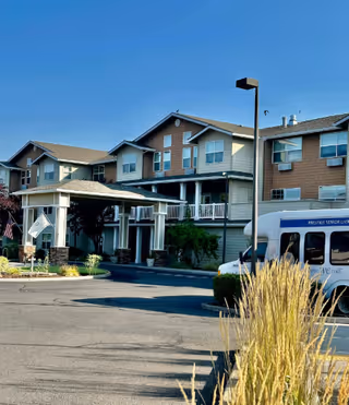Entrance of a three-story senior living building with a covered porte-cochere, parked shuttle van, and landscaped foreground under a clear blue sky.