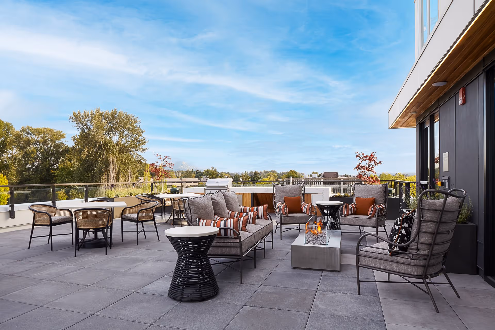 Rooftop patio with cushioned seating, a central fire pit, tables and chairs overlooking trees under a blue sky.