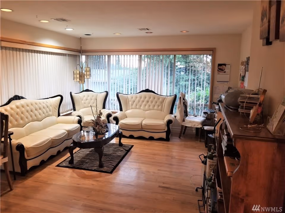 Bright living room with three white tufted sofas, a glass-top coffee table on a hardwood floor, and large sliding glass doors with vertical blinds.