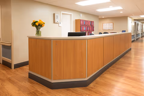 A reception desk area in a senior living facility with a wooden counter and a vase of yellow flowers on top. The floor is wood, and there are chairs and a bulletin board visible in the background along a hallway.