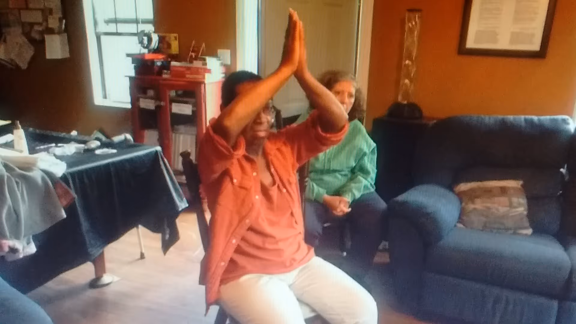 Two women sitting in a cozy living room with warm orange walls. One woman in an orange shirt and white pants is seated on a chair with her hands raised and pressed together above her head. The other woman in a green shirt is sitting on a dark blue couch with a patterned pillow. There is a table covered with a black cloth and various items on it, a window with blinds, and a red cabinet in the background.