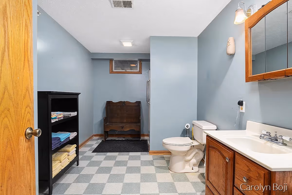 Bathroom with a toilet, sink and wooden vanity, shelving of folded towels and a wooden bench against blue-gray walls.