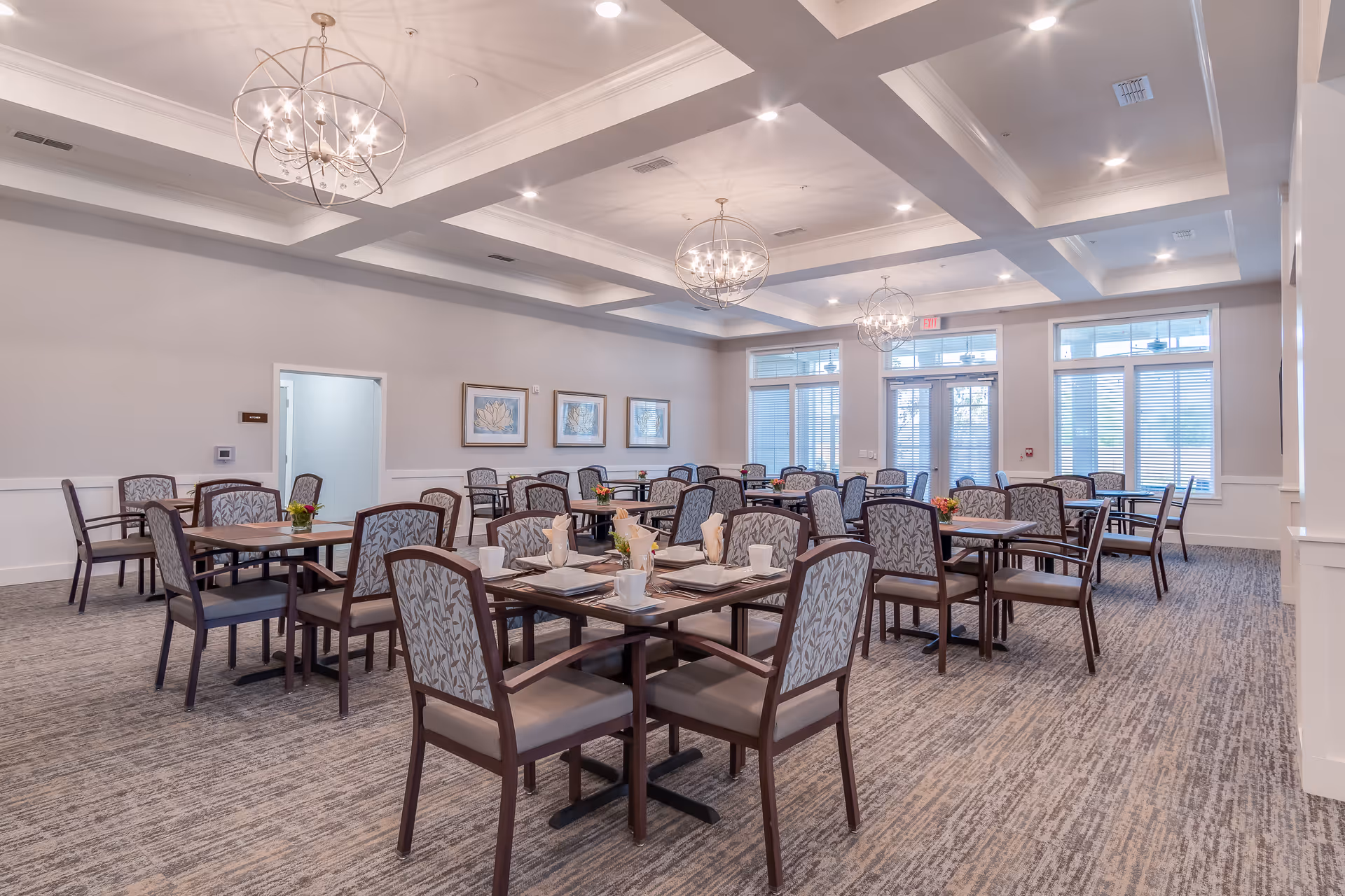 A spacious dining room with multiple tables and chairs arranged neatly. Each table is set with white plates, cups, and napkins. The room features large windows with blinds, three modern chandeliers hanging from a coffered ceiling, and framed artwork on the walls. The carpet is patterned in neutral tones, and the overall decor is elegant and inviting.
