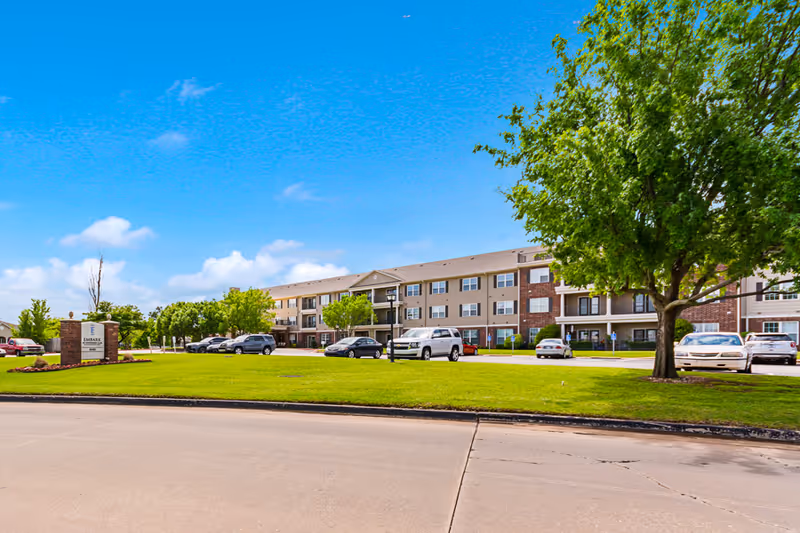 Exterior view of a three-story senior living facility building with a parking lot in front, green lawn, and trees under a bright blue sky with some clouds.