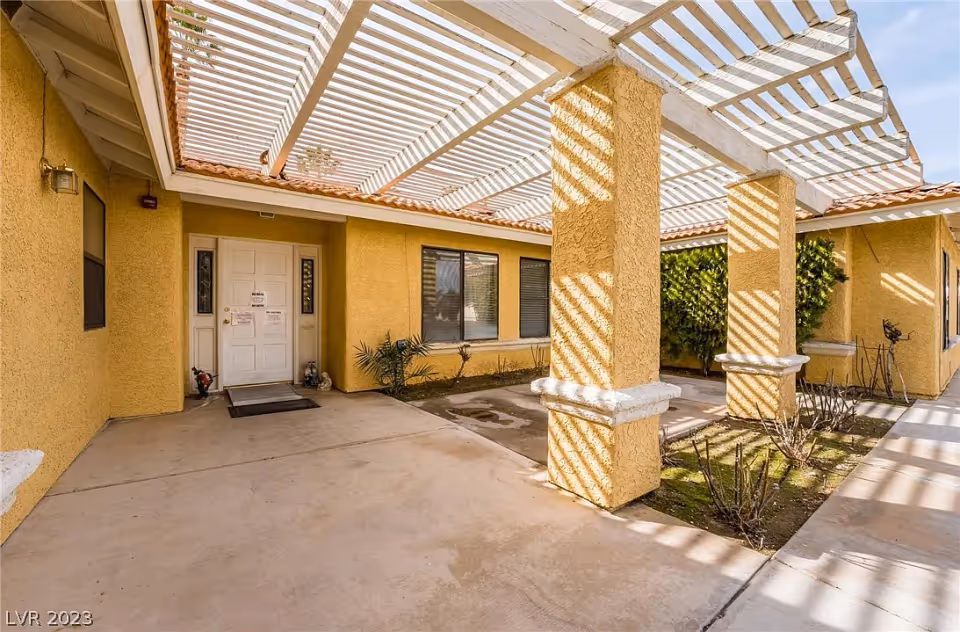 Outdoor covered walkway area with yellow stucco walls and white lattice roof casting striped shadows. There are two large columns, a white door with windows on either side, and some small plants along the building.