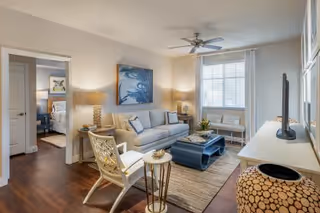 A bright and cozy living room with a white sofa, a blue coffee table, a white armchair, and a TV on a white stand. The room has wooden flooring, a ceiling fan, two table lamps, and a large window with white curtains. A doorway leads to a bedroom with a bed and artwork visible.