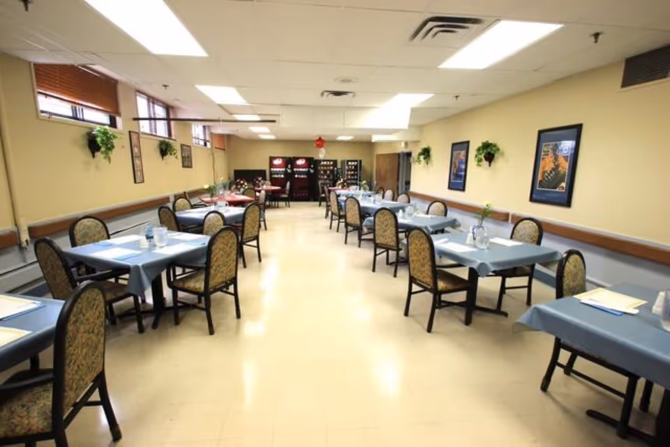 A dining room with several tables covered in blue tablecloths, each surrounded by chairs with patterned upholstery. The room has beige walls decorated with framed pictures and small wall-mounted plants. There are windows near the ceiling on the left side and vending machines at the far end of the room.
