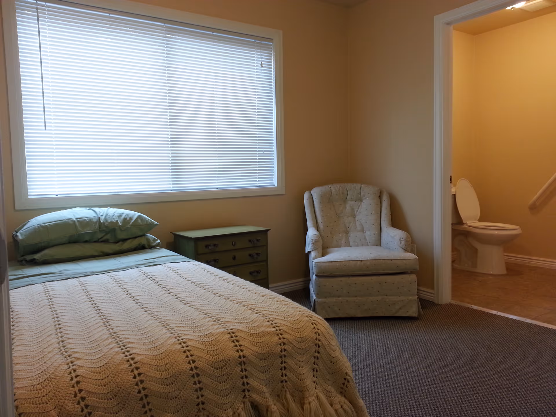 A small bedroom with a single bed covered with a beige knitted blanket and green pillows. Next to the bed is a small green nightstand with drawers. There is a cushioned armchair with a light patterned fabric near the entrance to a bathroom, where a toilet is visible. The room has beige walls and a large window with closed blinds.