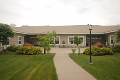 A single-story building with a beige exterior and a brown roof, surrounded by a well-maintained lawn and small trees. A concrete pathway leads to the entrance area, which has benches and flower beds with pink and yellow flowers. A black lamppost stands on the right side of the pathway.