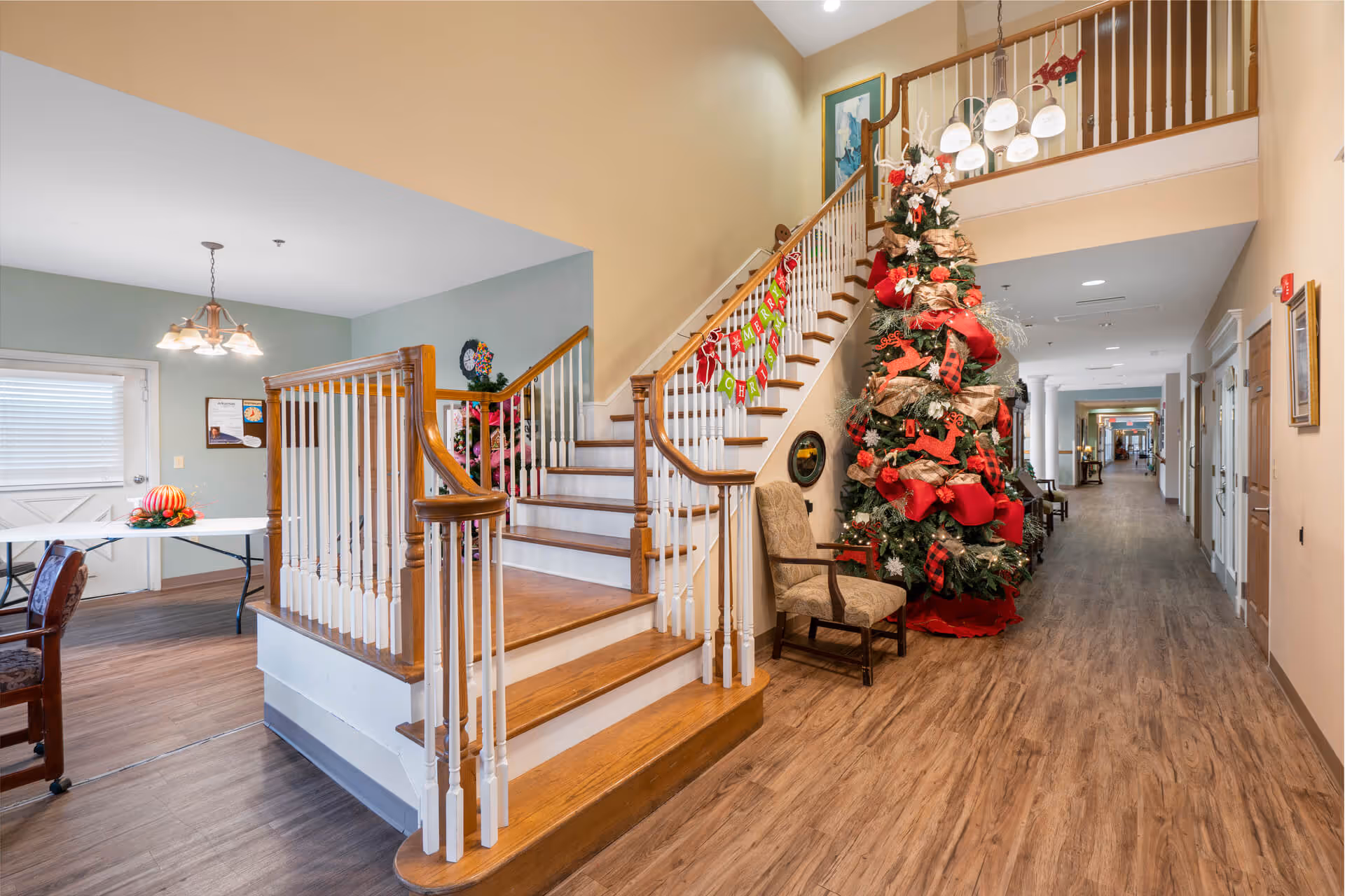 Interior view of a senior living facility hallway with a wooden staircase decorated for Christmas with a large Christmas tree adorned with red ribbons and ornaments. There is a beige armchair next to the tree and a long corridor with doors and chairs along the walls. A small table with chairs is visible to the left under a hanging light fixture.