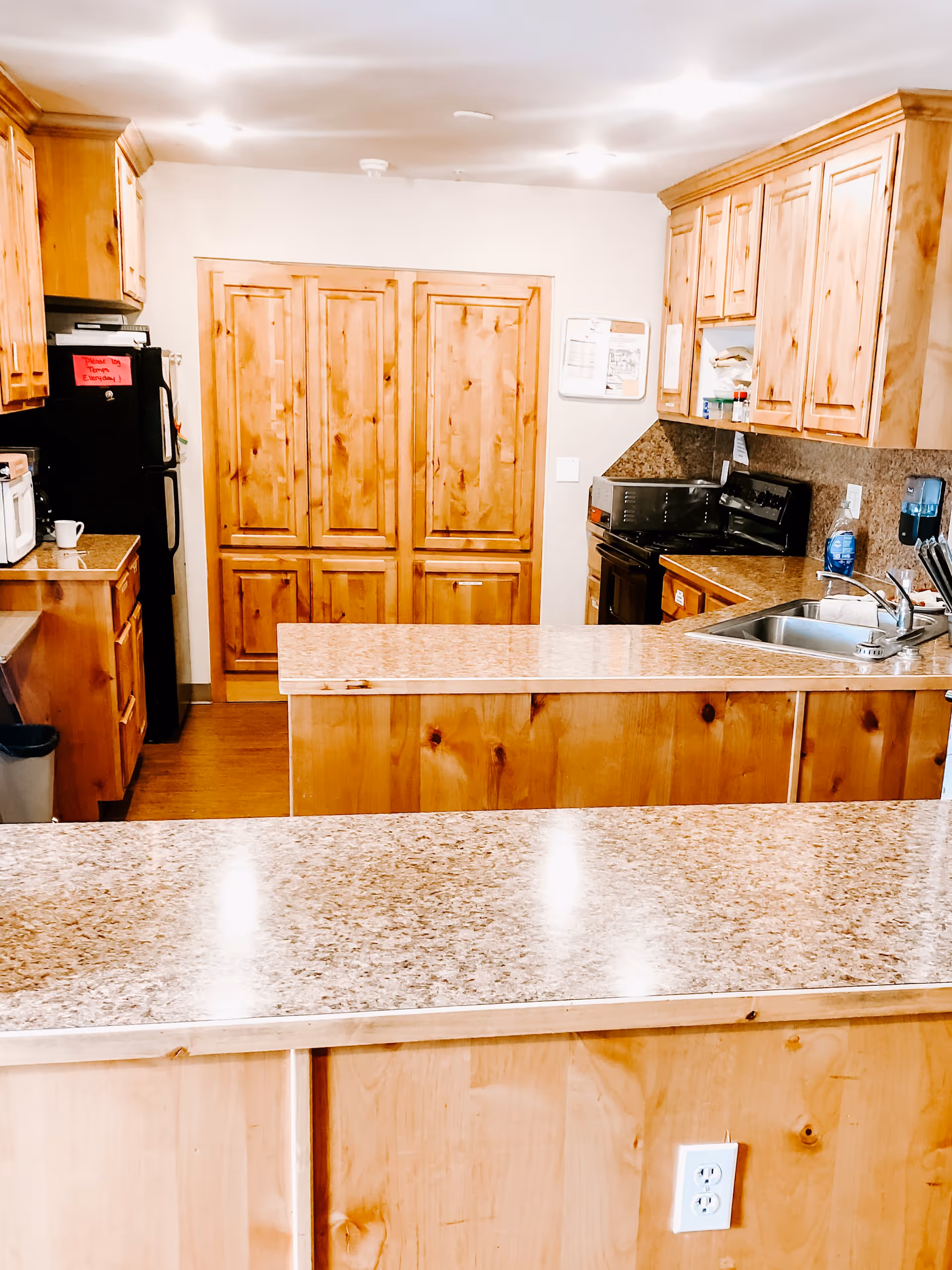 A kitchen area with wooden cabinets and drawers, granite countertops, a black refrigerator with a red note on it, a microwave, a stove, a sink, and various kitchen items. The walls are light-colored and the floor is wooden.