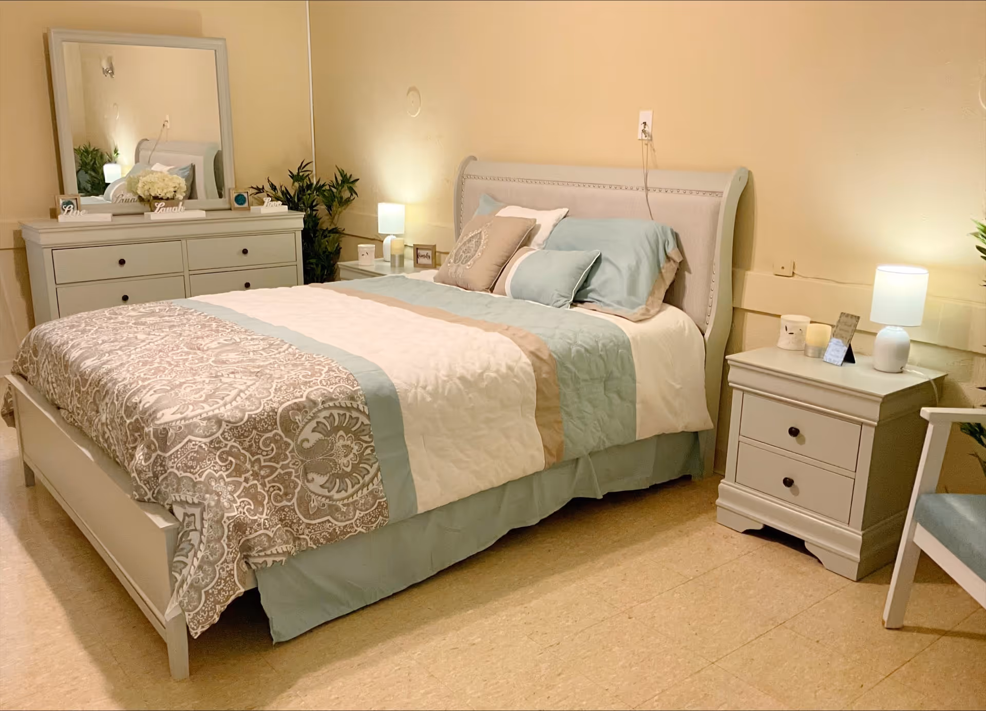 A cozy bedroom with a neatly made bed featuring a patterned comforter and multiple pillows. There is a white dresser with a large mirror above it, decorated with small plants and decorative signs. A nightstand with a lamp and candles is beside the bed, and a chair is partially visible on the right. The room has soft lighting and a warm, inviting atmosphere.