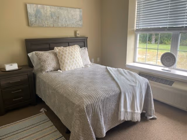 A neatly made bed with a gray quilted bedspread and three decorative pillows in a bedroom. There is a dark wooden nightstand with a white clock on it to the left of the bed. A large window with blinds is on the right side, letting in natural light and showing greenery outside. A light-colored rug is partially visible on the floor near the bed.