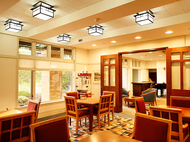 Bright communal dining area with wooden tables and chairs, patterned rug, large windows, and a connected seating lounge visible through glass-paneled double doors.
