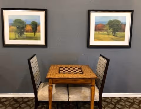 A small wooden table with a built-in chessboard and two upholstered chairs positioned on either side, set against a gray wall with two framed landscape paintings hanging above.