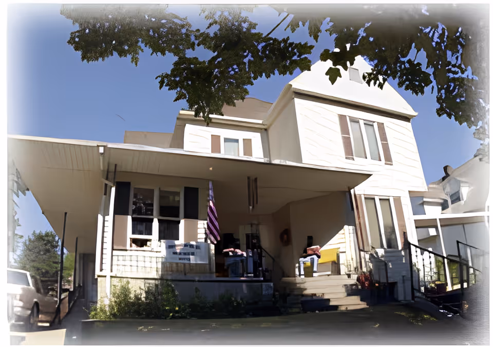 Exterior view of a two-story residential building with a covered front porch, steps leading up to the entrance, and an American flag hanging near the door. The building is surrounded by trees and has a clear blue sky in the background.