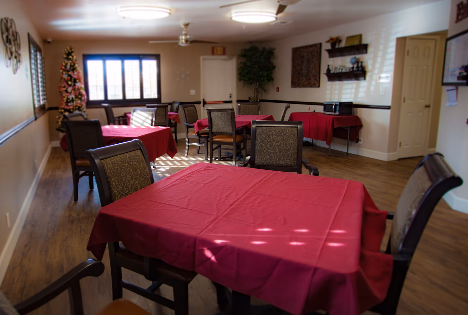 Dining room with several tables covered in red tablecloths and chairs and a decorated Christmas tree by the window.