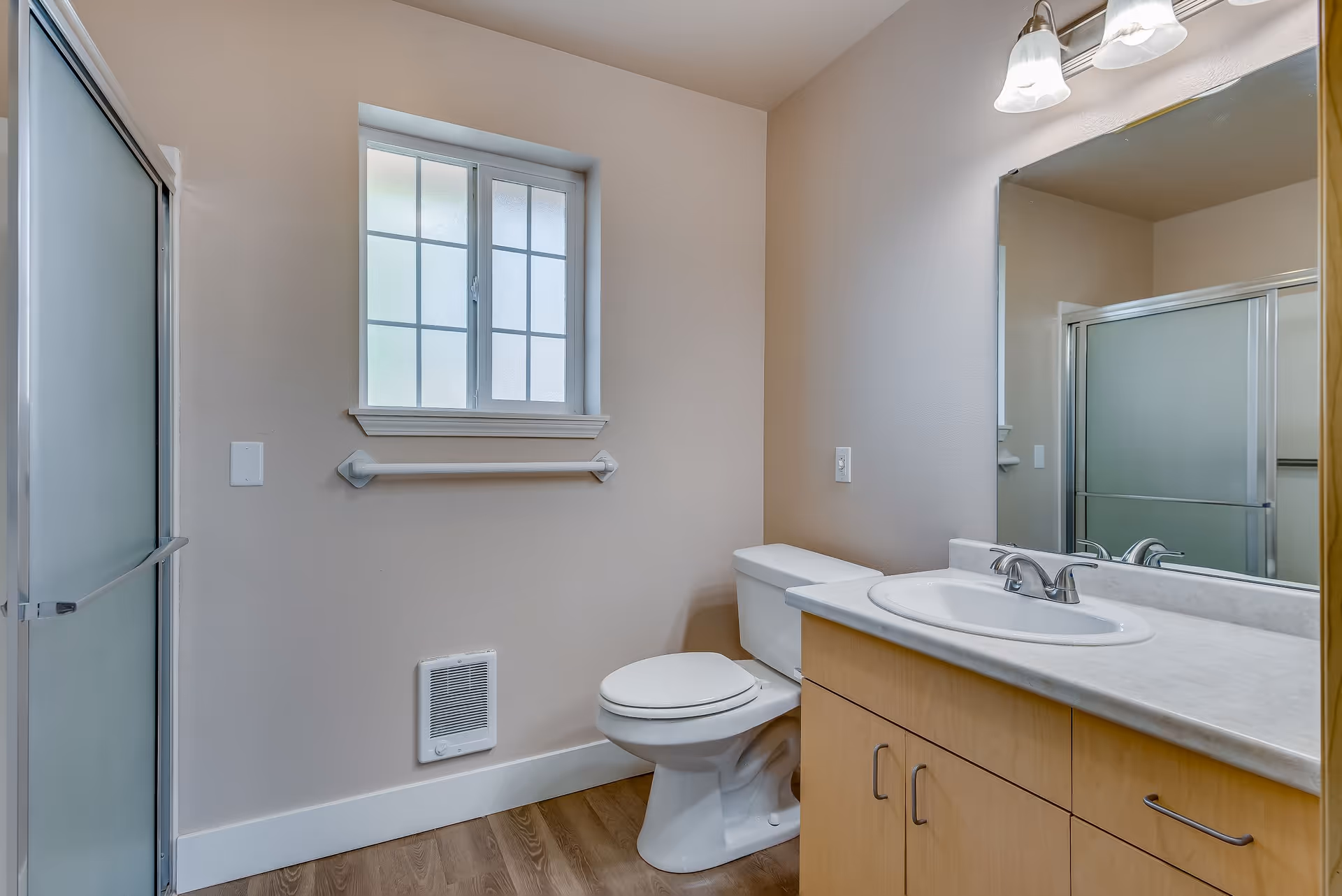 A clean and modern bathroom featuring a toilet, a wooden vanity with a white sink and countertop, a large mirror above the sink, a frosted glass shower door, a window with frosted glass panes, a towel bar, and light fixtures above the mirror. The floor is wood-style and the walls are painted beige.