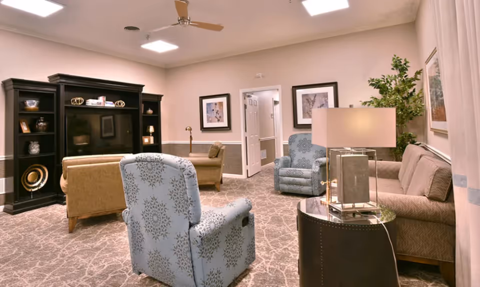 A cozy living room in Dogwood Bend featuring a beige sofa, two patterned armchairs, a black entertainment center with a large TV, decorative shelves, framed artwork on the walls, a floor lamp, a table lamp on a round side table, and a potted plant in the corner.