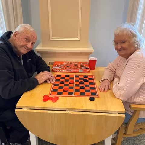 Two elderly individuals sitting at a wooden table playing a game of checkers. The man on the left is wearing a black jacket, and the woman on the right is wearing a light pink sweater. A red cup and the checkers game box are on the table. They are in a room with light-colored walls and a decorative column behind them.