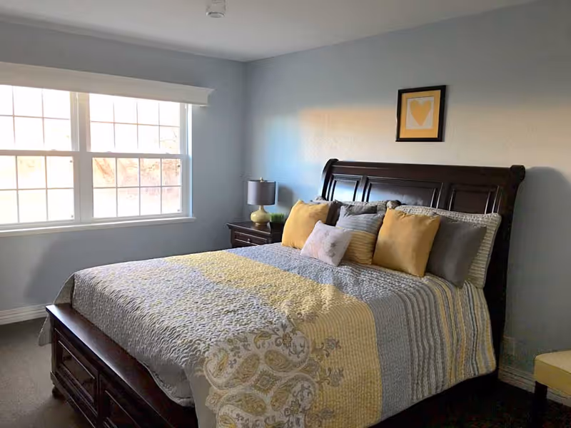 Sunlit bedroom with a large window, a dark wood bed made with yellow and gray bedding, a nightstand with a lamp, and framed heart artwork on the wall.