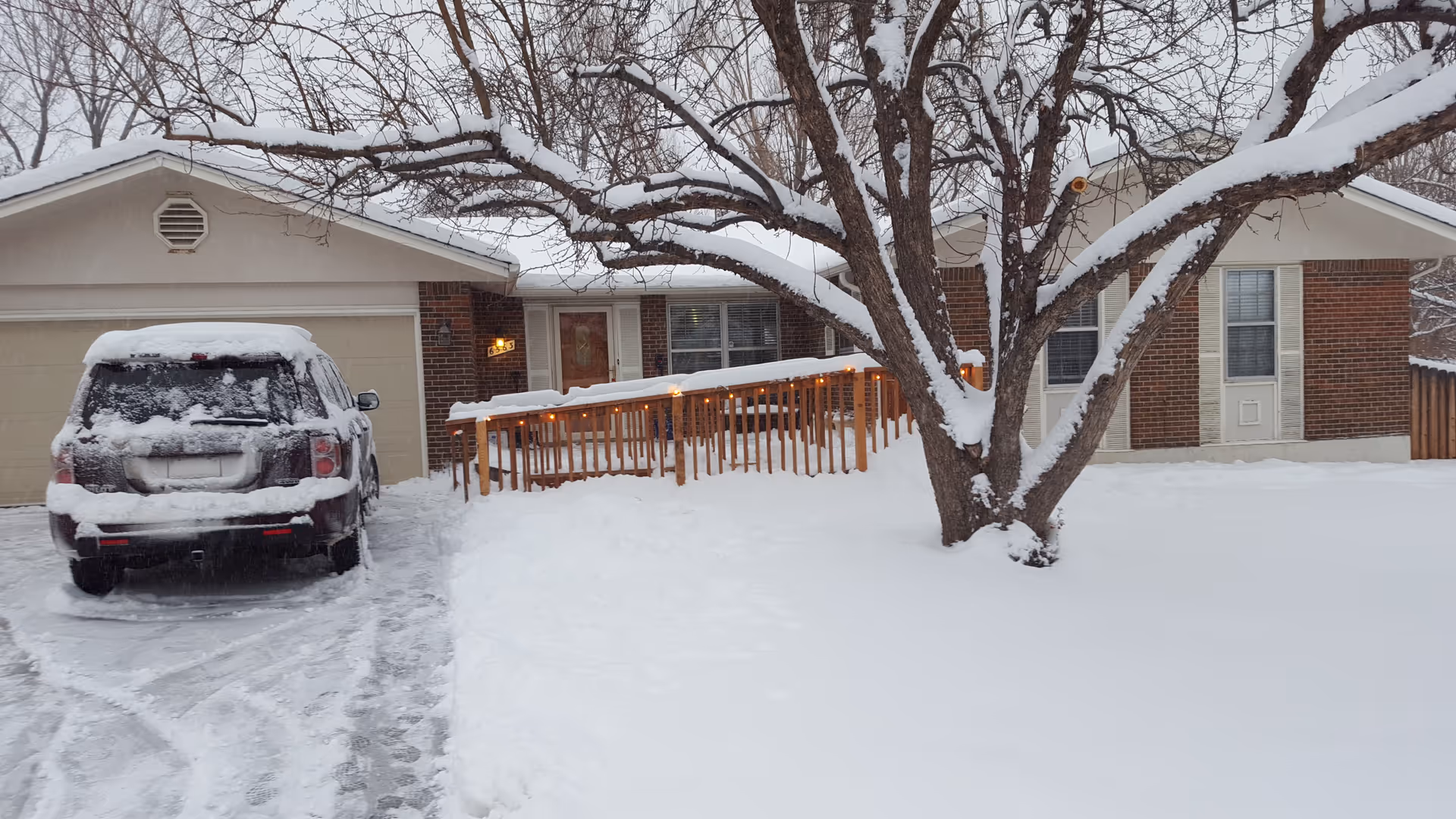 A single-story brick house covered in snow with a snow-covered black SUV parked in the driveway. A large tree with snow on its branches stands in the front yard, and a wooden ramp with string lights leads to the front door. The ground is completely covered in snow.