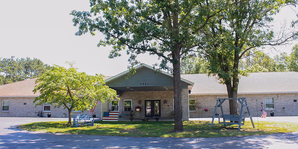 Front exterior view of a single-story senior living facility building with a covered entrance, surrounded by trees and green lawn. There are benches and a swing on the lawn, along with American flags and decorative elements.
