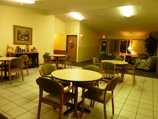 Interior view of a senior living facility common area with round tables and chairs arranged on a tiled floor. In the background, there is a seating area with patterned armchairs, a lamp, and a large plant. The walls are light-colored, and there is a framed painting and decorative items on a side table.
