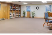 A spacious room with carpeted floor, wood-paneled wall with built-in bookshelves filled with books, two framed pictures and a clock on a white wall, two potted plants, and a wooden table with blue cushioned chairs partially visible on the right.