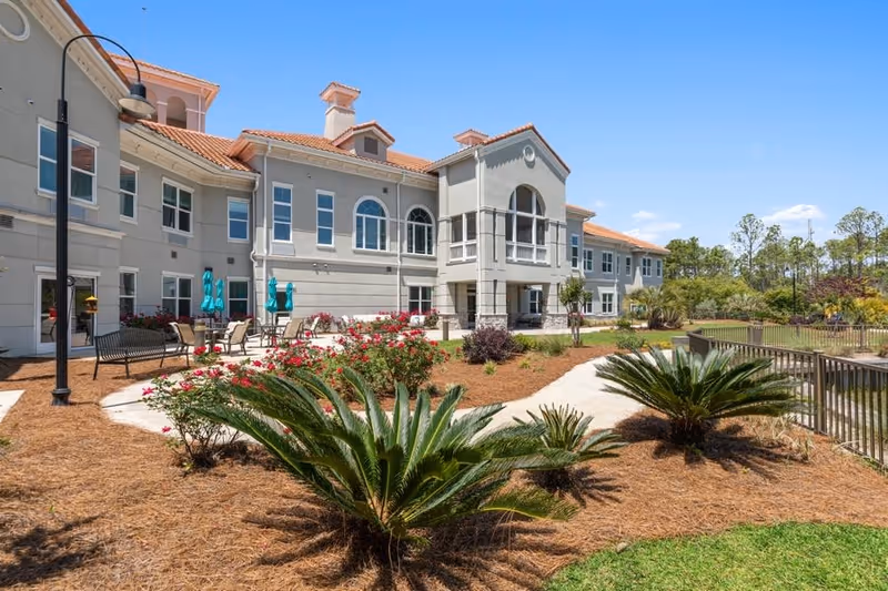 Exterior view of a two-story senior living facility building with beige walls and a red-tiled roof. The landscaped garden in front features palm plants, flowering bushes, and a paved walkway. Outdoor seating with tables and turquoise umbrellas is visible near the building. The sky is clear and blue.
