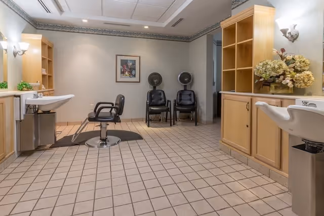 Interior view of a salon area in a retirement community featuring two black salon chairs in front of sinks, two black chairs with hair dryers behind them, light-colored tiled floor, wooden cabinetry, and a framed picture on the wall.