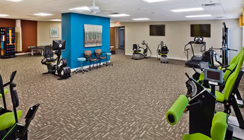 A spacious senior living facility exercise room with various fitness equipment including stationary bikes and weight machines. The room has carpeted flooring, a blue accent wall with framed artwork, several chairs lined up against the wall, and two large flat-screen TVs mounted on the far wall.