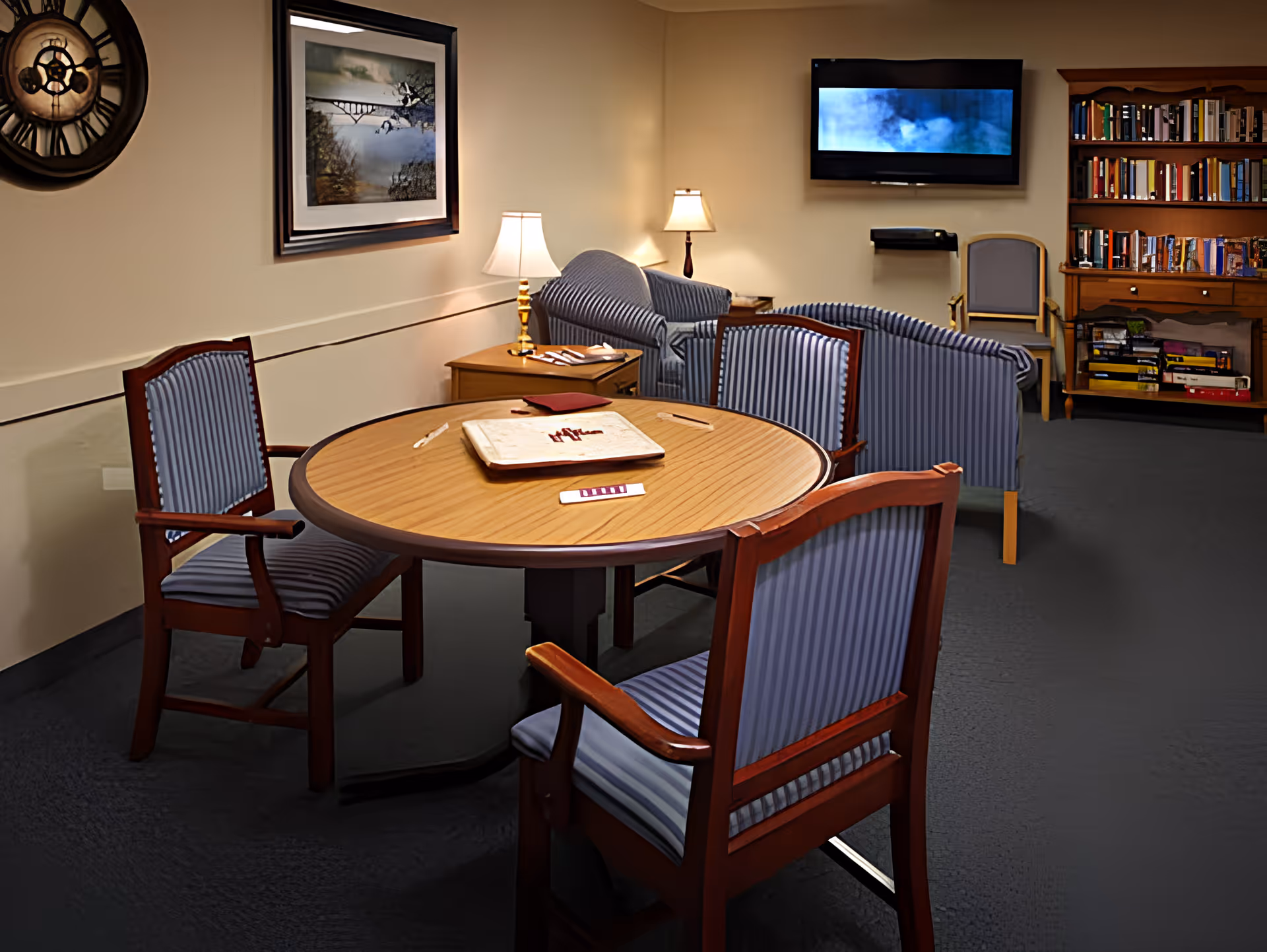 A cozy common room with a round wooden table surrounded by four striped upholstered chairs. In the background, there are two striped armchairs, a side table with two lamps, a wall-mounted TV, a wooden bookshelf filled with books and games, and a framed picture on the wall.