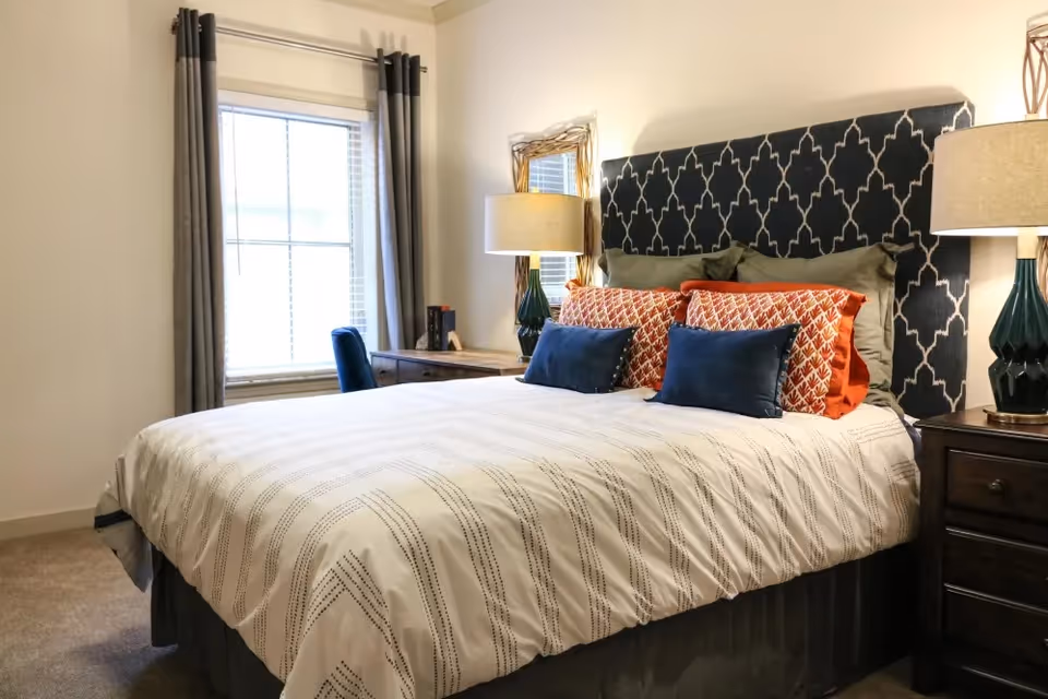 A neatly made bed with a patterned dark blue headboard, white and gray striped bedding, and multiple decorative pillows in orange and blue. There are two bedside tables with matching lamps, a window with gray curtains, and a wooden desk with a blue chair in the background.