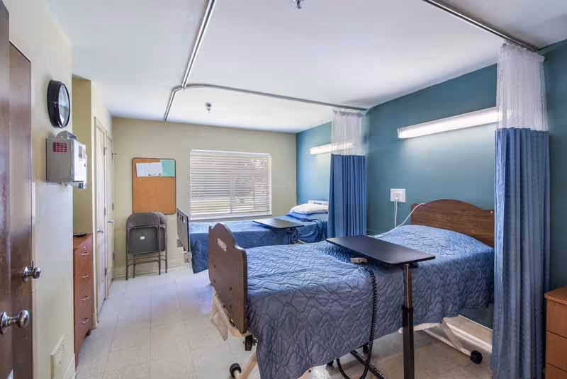 A room with two hospital-style beds separated by a curtain. Each bed has a blue quilted blanket and an adjustable overbed table. The walls are painted blue and beige, and there is a window with blinds letting in natural light. A clock and a bulletin board are visible on the wall near the door.