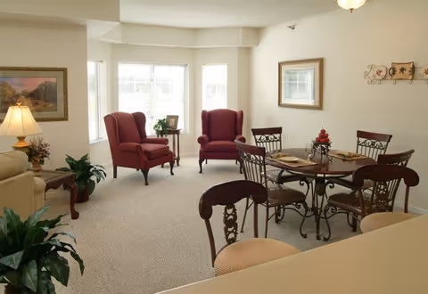 A cozy living area featuring two red upholstered armchairs near a bay window with a small side table between them. There is a round dining table with four chairs, set with placemats and a centerpiece. The room has beige carpet and walls decorated with framed artwork and a wall-mounted decorative piece. A lamp and some plants add to the warm ambiance.