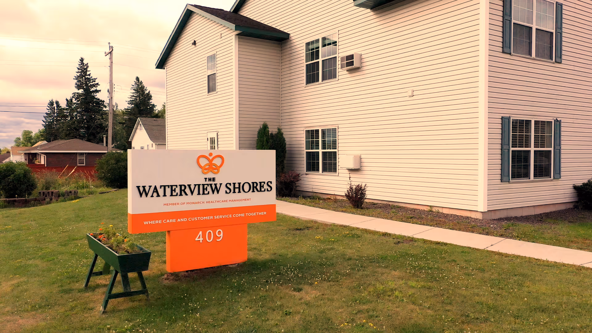 Exterior view of The Waterview Shores facility showing a white building with multiple windows and a sign in front on a grassy lawn. The sign displays the facility name, logo, and address number 409. There are trees and other houses visible in the background under a cloudy sky.