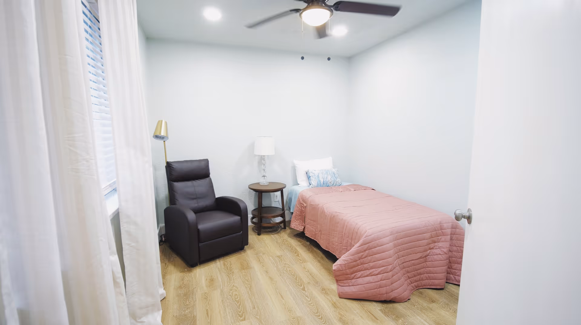 A small, simple bedroom with light-colored walls and wood flooring. The room contains a single bed with a pink quilt and white pillow, a dark brown recliner chair, a round wooden side table with a white lamp, and a standing lamp near the window with white curtains. A ceiling fan with a light is mounted on the ceiling.