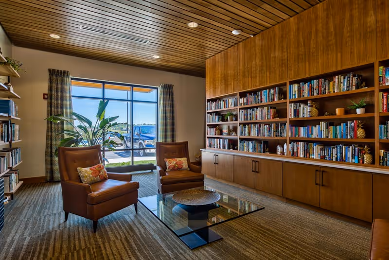 A cozy senior living facility library or reading room with two brown leather armchairs, each with a floral pillow, positioned around a glass coffee table. The room features a large wooden bookshelf filled with books and decorative items, a large window with patterned curtains letting in natural light, and a wooden ceiling.