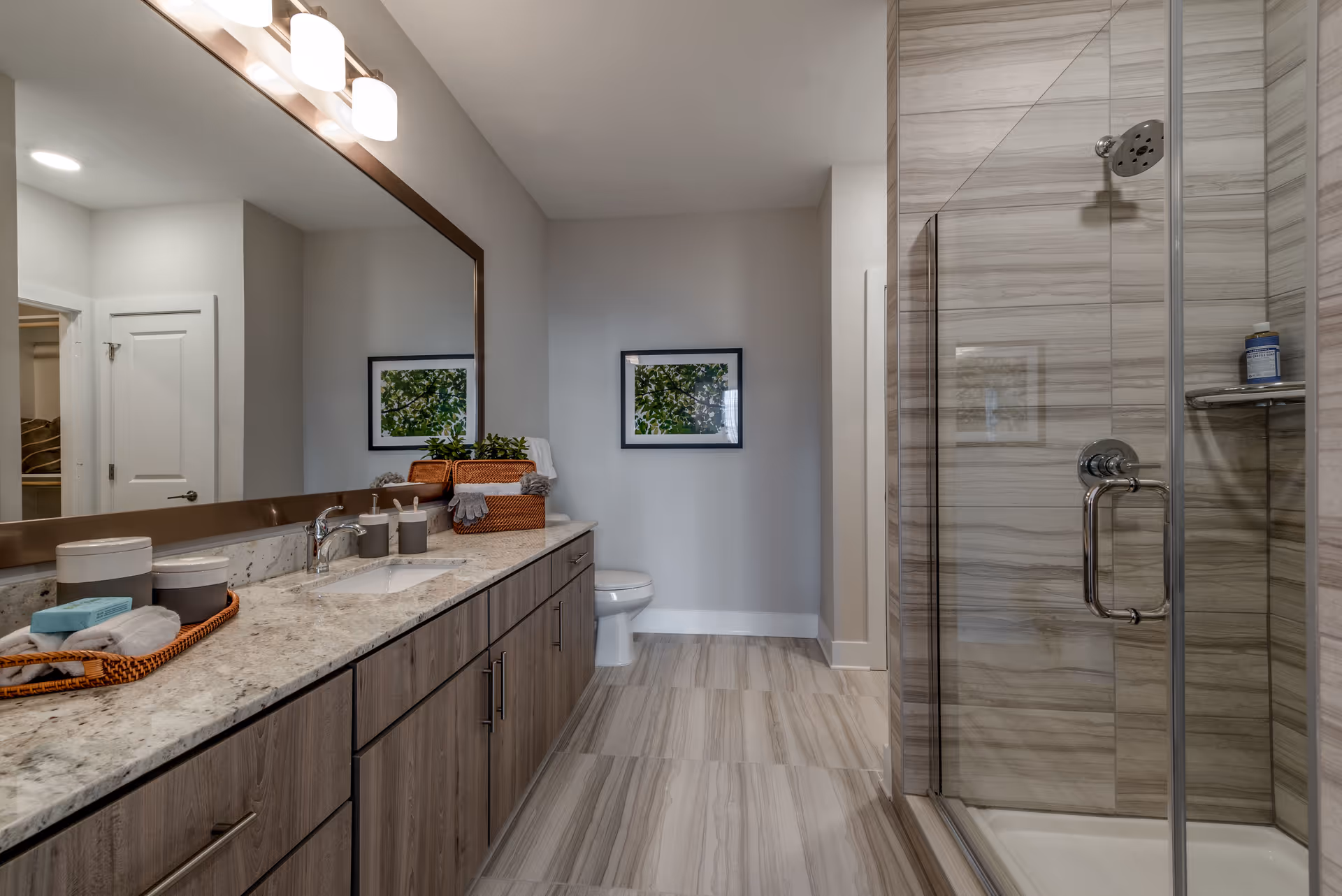 Modern bathroom with a large mirror above a granite countertop with a sink, wooden cabinets below, a glass-enclosed shower with tiled walls, a toilet, and a framed picture on the wall.