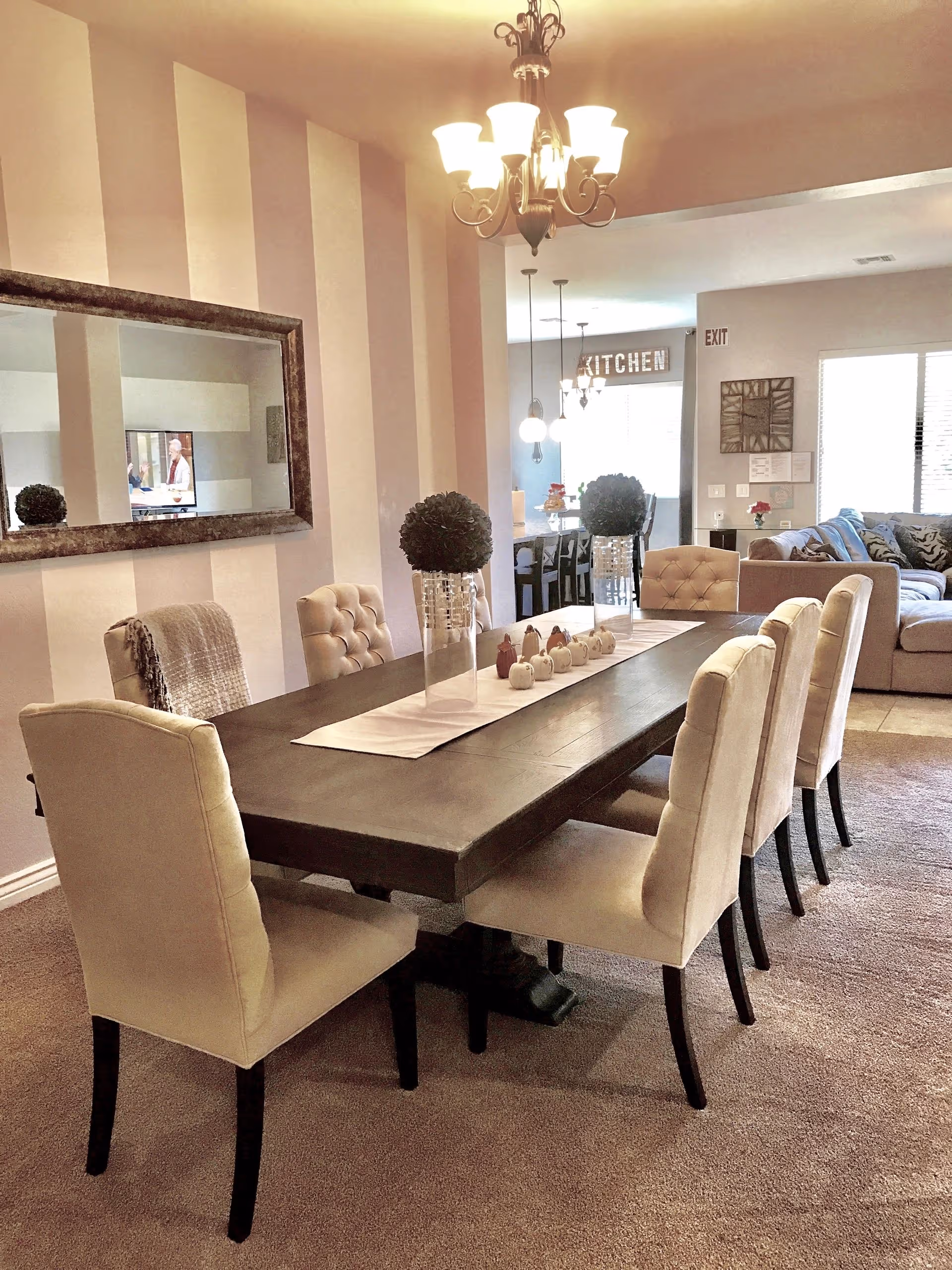 A dining room with a dark wooden rectangular table surrounded by eight beige upholstered chairs. The table is decorated with a white runner, two glass vases with green topiary balls, and small decorative pumpkins. A large mirror hangs on the striped wall to the left. In the background, a living room with a beige sectional sofa and a kitchen area with pendant lights and a sign that reads 'KITCHEN' are visible. A chandelier hangs above the dining table.