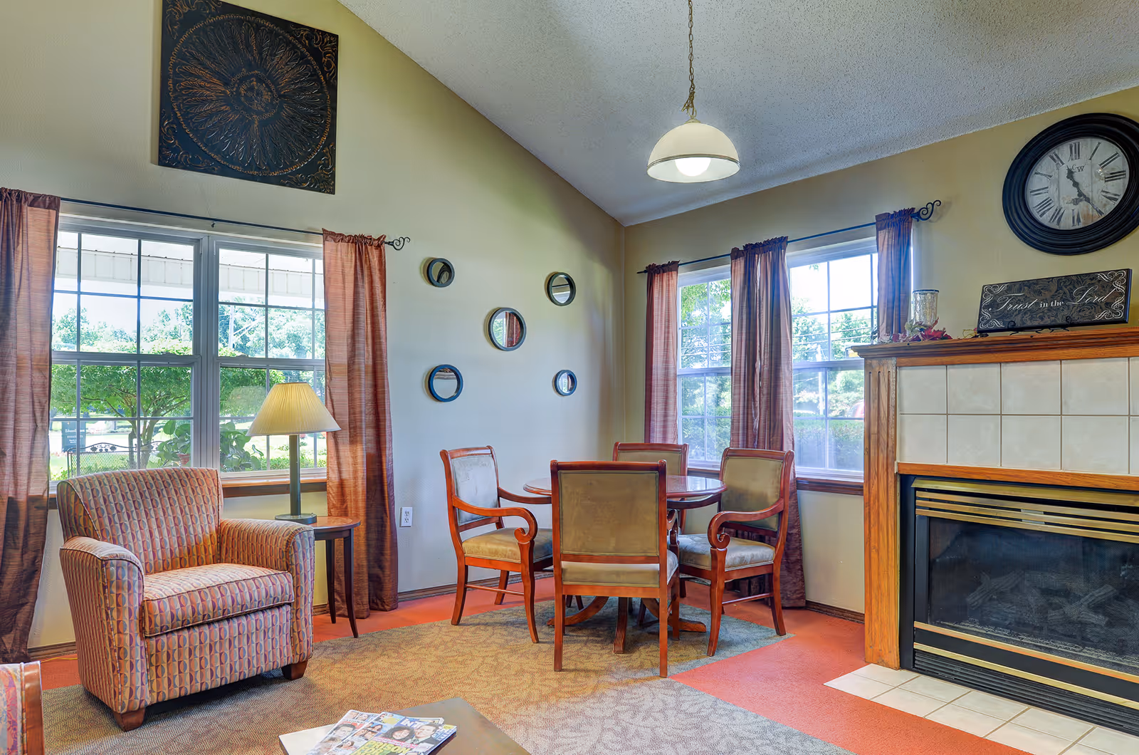 A cozy living room area with a patterned armchair, a round wooden table surrounded by four chairs, a fireplace with a clock and decorative sign above it, and two large windows with brown curtains letting in natural light. The walls are light-colored and decorated with small round mirrors and a large decorative piece above one window.