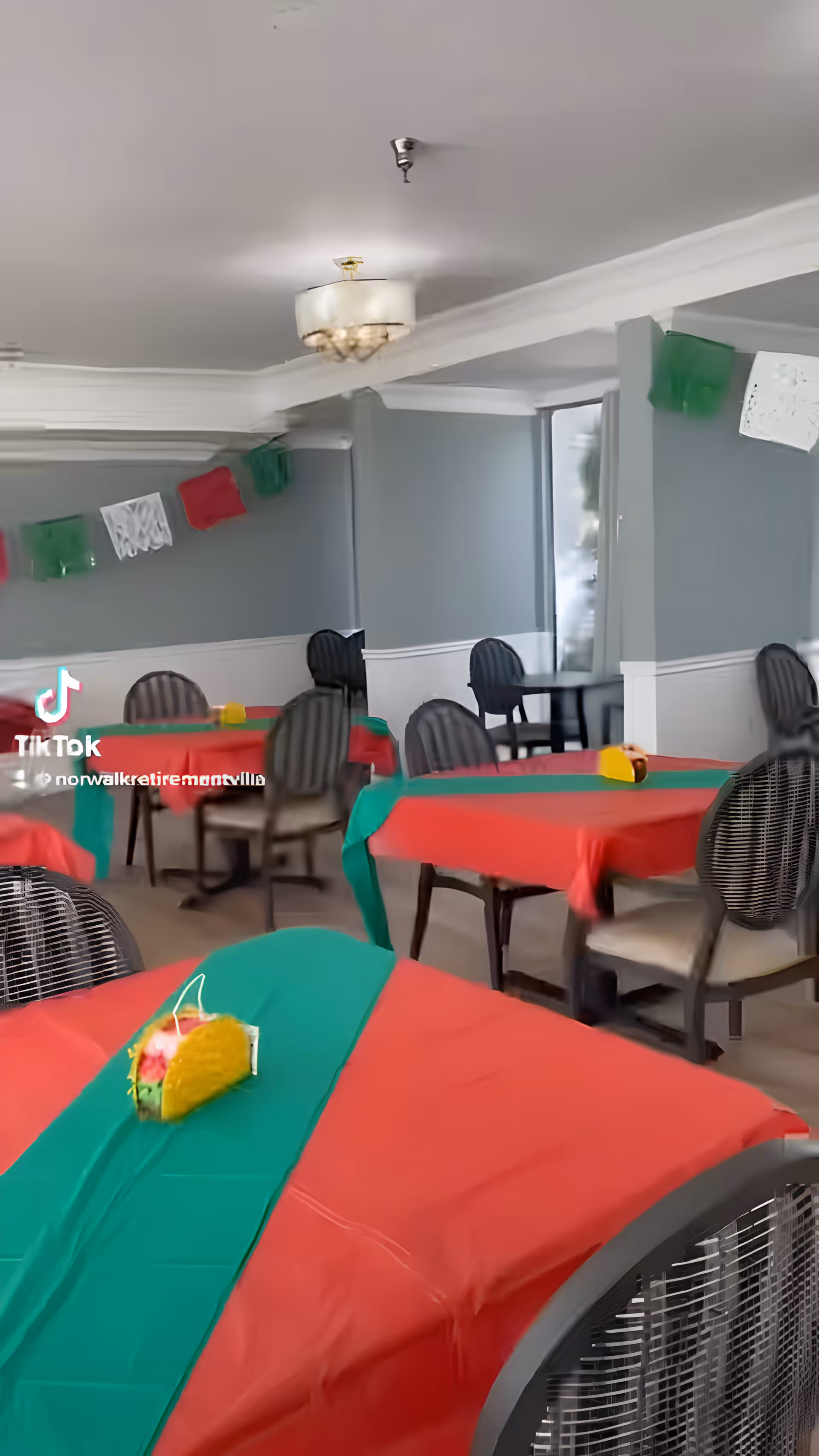 Dining room with multiple tables covered in red and green tablecloths and chairs, decorated with hanging paper banners.