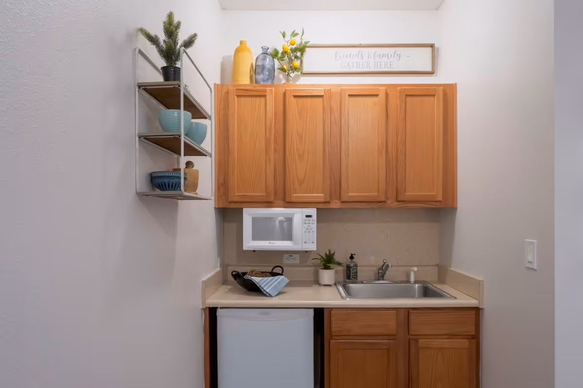 Small kitchenette with wooden cabinets, a microwave over the counter, sink, mini fridge and open shelving with dishware and a decorative sign above.