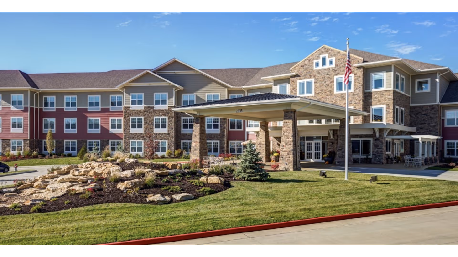 Exterior view of Cedarhurst of Columbia, a large multi-story senior living facility with a covered entrance supported by stone pillars, landscaped garden with rocks and small trees, an American flag on a flagpole, and a clear blue sky.