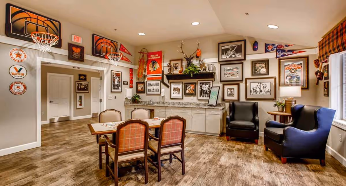 A cozy recreational room with a wooden floor, a table with four chairs in the center, and two black armchairs near a window with plaid curtains. The walls are decorated with numerous framed sports memorabilia, including basketball hoops, pennants, trophies, and photographs. There is a countertop with cabinets underneath along one wall.