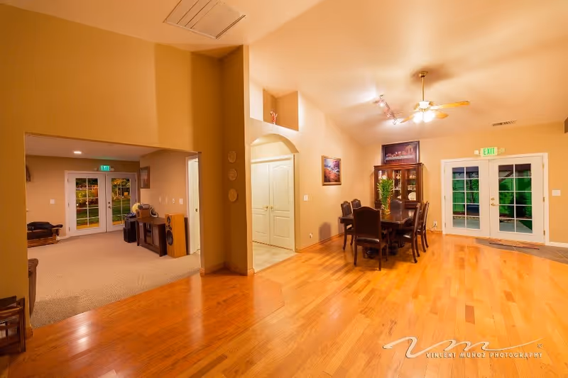 Interior view of a senior living facility showing a dining area with a wooden table and six chairs, a china cabinet, and two sets of glass double doors leading outside. The room has hardwood floors, beige walls, ceiling fan with lights, and framed artwork on the walls. Adjacent to the dining area is a carpeted living space with a bench and speakers, visible through an open doorway.
