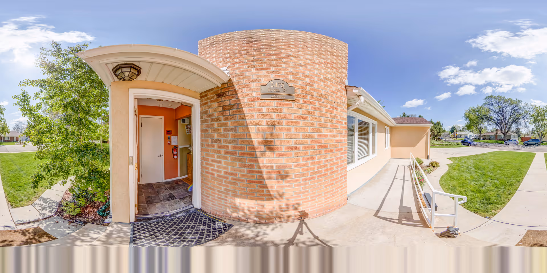 Exterior front entrance of a brick-clad assisted living/memory care home with an open doorway, ramp, and landscaped lawn under a blue sky.