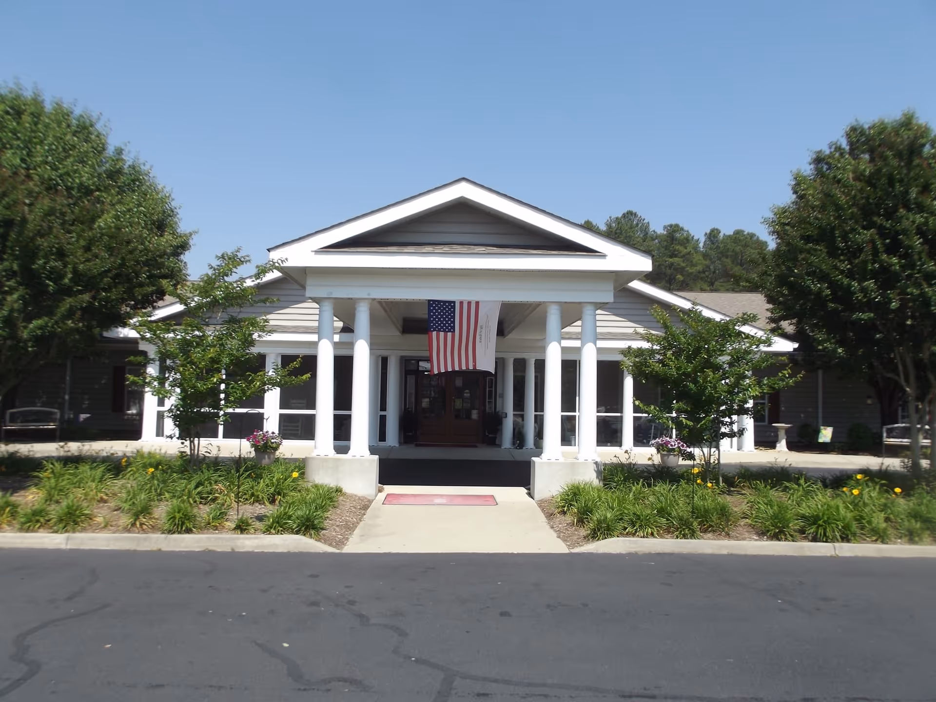 Front exterior view of Commonwealth Senior Living at Kilmarnock building with white columns, an American flag hanging under the entrance roof, surrounded by greenery and trees on a sunny day.