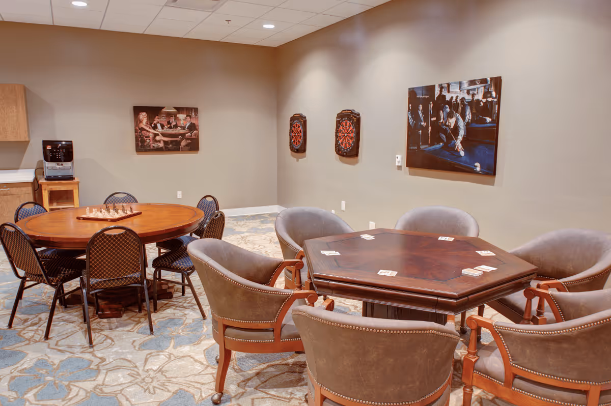 A recreational room with two tables and chairs. One table is round with a chessboard on it and six chairs around it. The other table is hexagonal with playing cards on it and six upholstered chairs around it. Two dartboards are mounted on the wall, along with two framed pictures depicting people playing games. The room has a patterned carpet and neutral-colored walls.